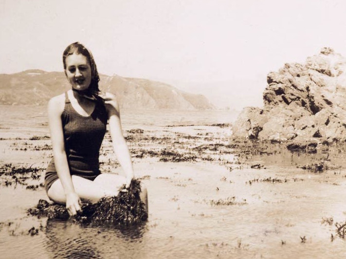 First seaweed field trip to Island Bay, January 1943, Wellington, maker unknown. Purchased 2007. Te Papa (CA000903/001/0003/0001) A sepia image of a woman in a bathing suit sitting next to some seaweed on a beach.