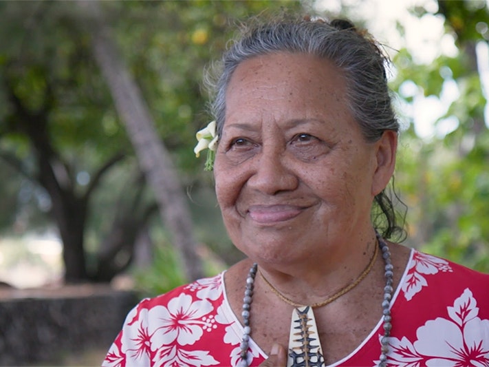 Still from Sarah Vaki interview. Te Papa Head and shoulders of a woman with a floral dress and a flower behind her right ear smiling at someone off camera.