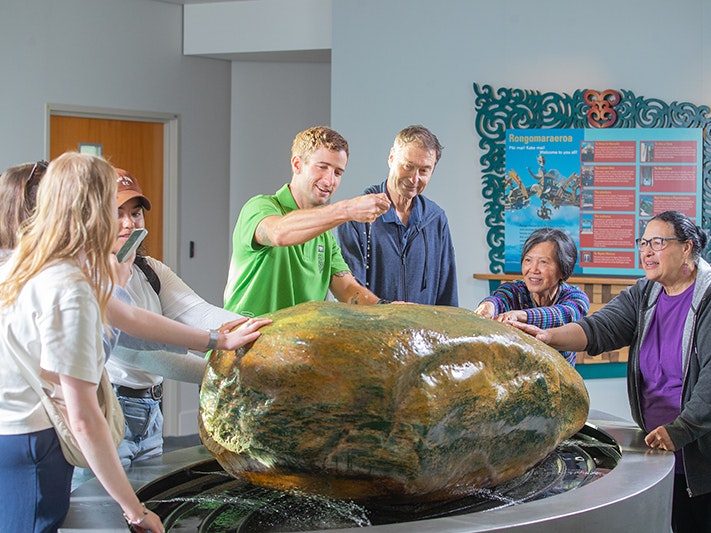 Te Papa Tours. Photo by Jeff McEwan, 2025. Te Papa (267107) A man in a bright green shirt is sprinkling small stones on a large piece of pounamu that has water spraying on it. He is surrounded by six other people looking on with some of them touching the stone.
