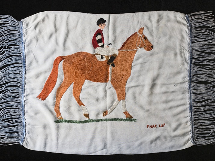 Hand embroidered cushion cover of Phar Lap and jockey, Melbourne Cup 1930. 1995.0032.0001. Copyright National Museum of Australia / CC BY-SA 4.0. A piece of embroidery with a jockey sitting on a horse and the words PHAR LAP stitched on the bottom-right corner.