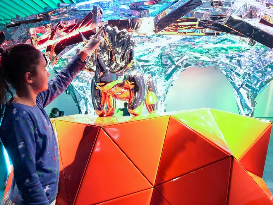 Child in the Curious Creatures and Marvellous Monsters exhibition with artwork by Gregor Kregar, Anthropocene Shelter, 2018, mylar, recycled timber, neon. Site-specific artist project, 2018. Te Papa Child in a brightly coloured environment touching a silver artwork model.