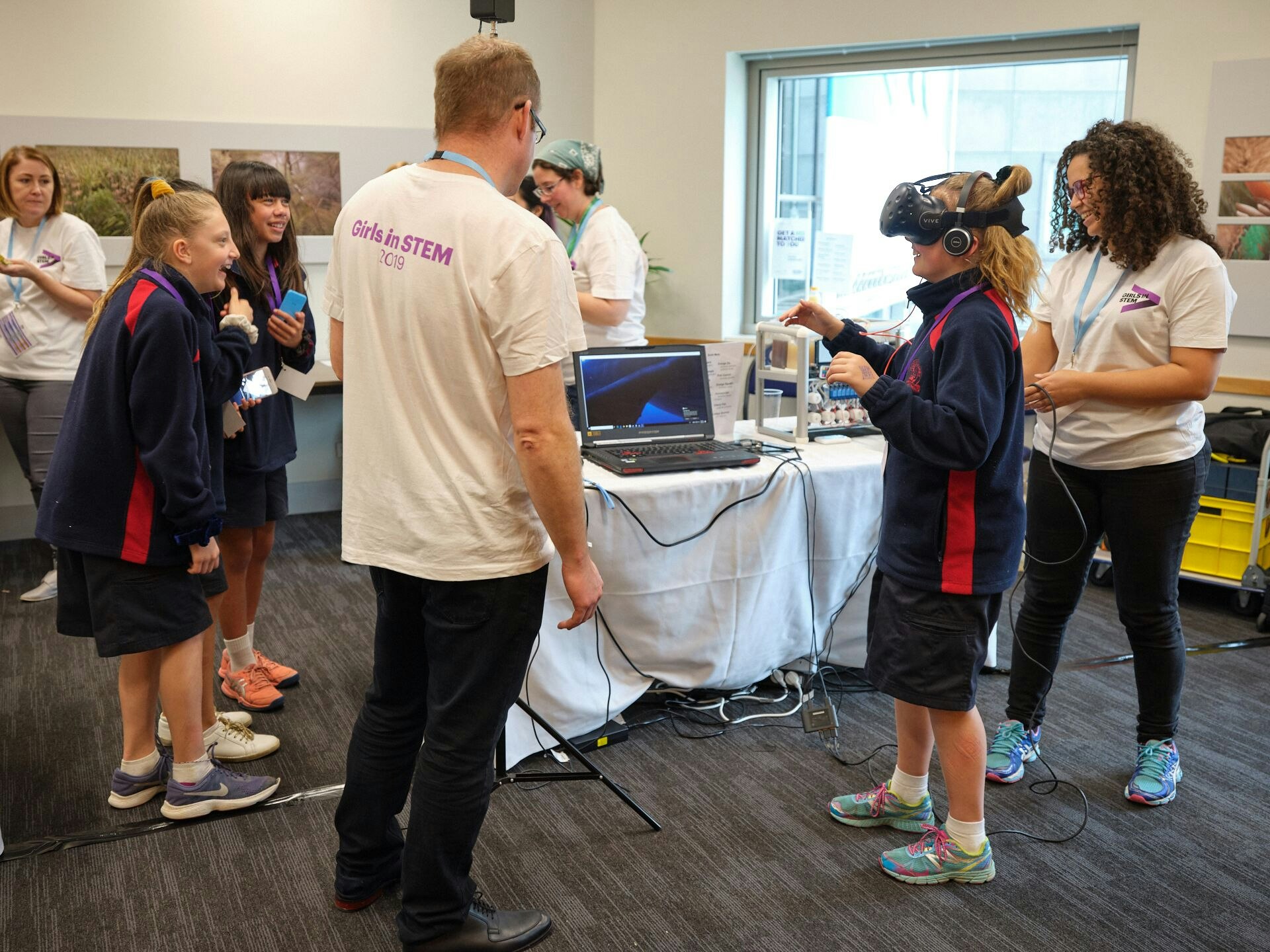 Girls having a VR experience. 2019. Photo courtesy of Accenture Adults and school children in a room. One child has a VR headset on.