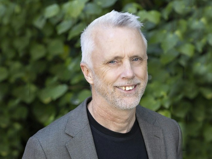 Phil Edgar, 2026. Photo by Michael O'Neill. Te Papa Head and shoulders of a man smiling at the camera. He is standing outside in front of a kawakawa hedge.