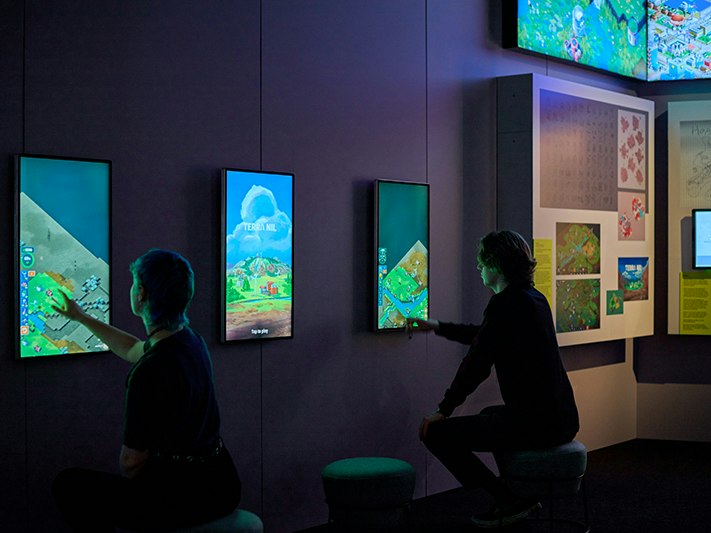 Game Worlds, 2025, ACMI. Photo © Eugene Hyland. Two people are in a darkened gallery. Both are pressing on digital screens mounted on the wall in front of them.