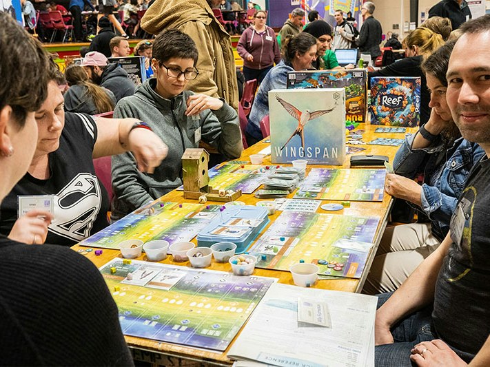 Image courtesy of Wellycon A group of people sit in a crowded room around a table playing the board game Wingspan